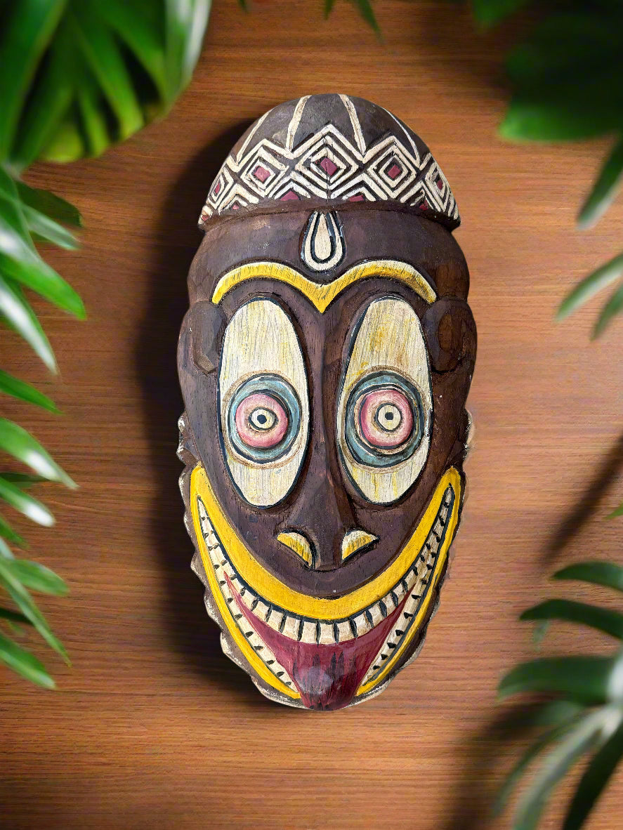 Wooden tribal mask with geometric crown, bulging eyes, and a wide yellow smile on a wooden surface